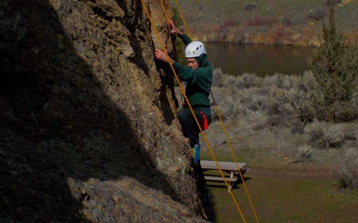 A person wearing safety gear is secured by ropes as they climb a rock wall. There is a river in the background.